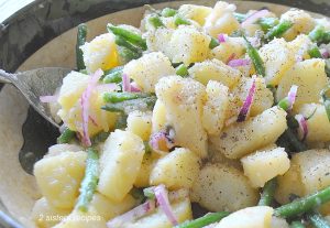 Chunks of boiled potatoes, and greens beans are tossed in a large salad bowl with seasonings and sliced red onions.