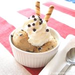 A white ramekin filled with of coffee flavored ice cream with whipped cream, chocolate chips and 2 swirls cookies on top.
