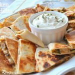 A white platter filled with pieces of quesadillas filled with chicken, mushrooms and onions, with sour cream in a white bowl in the center.