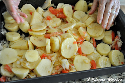 A large black roasting pan filled with raw sliced potatoes, tomatoes and seasonings.