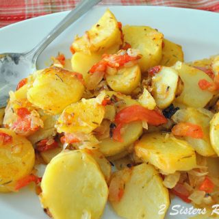 A white serving platter filled with roasted sliced potatoes and bits of tomatoes with a serving spoon.