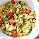 A white salad bowl filled with a mixture of tortellini pasta, cheese tomatoes and fresh basil.