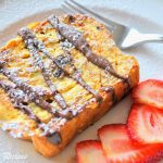 A Slice of French Toast served on a white plate with freshly sliced strawberries on the side.