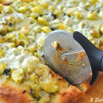 A pizza cutter slicing the pizza topped with chopped zucchini.