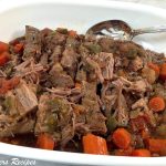 A white serving platter filled with pieces of tender brisket, chopped carrots and gravy.