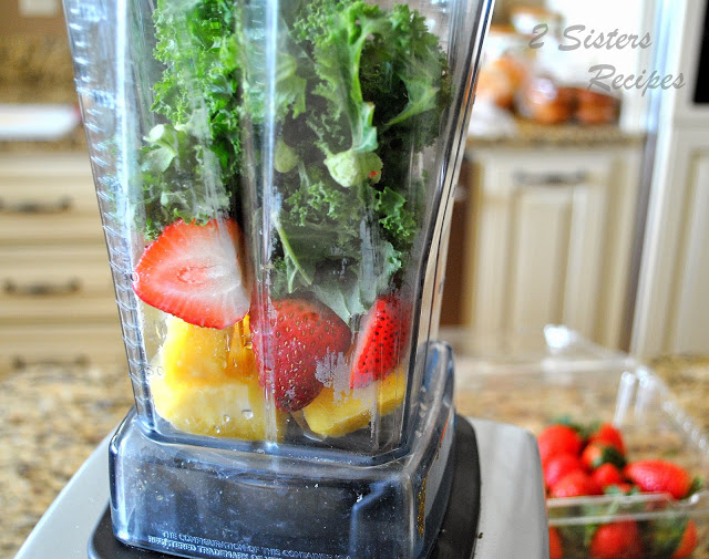 A blender filled with fresh strawberries, chunks of pineapples and kale.