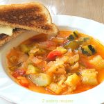 A white bowl of soup loaded with chunks of tomatoes and zucchini with a slice of toast on the side.
