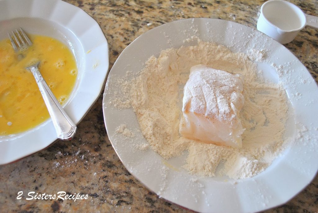A small piece of raw cod is coated in flour in a white plate. by 2sistersrecipes.com 