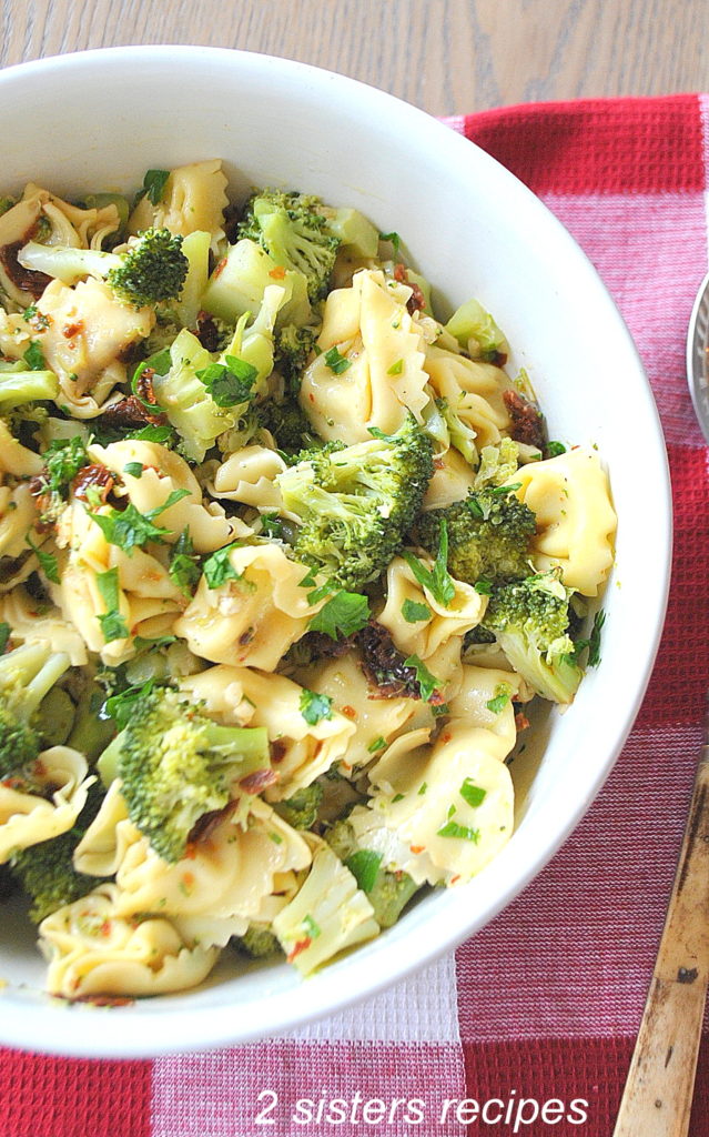 A white salad bowl filled with Tortellini and Broccoli salad on a red and white table cloth. 