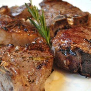 4 lamb chops grilled and placed on a white plate with fresh rosemary in the center.