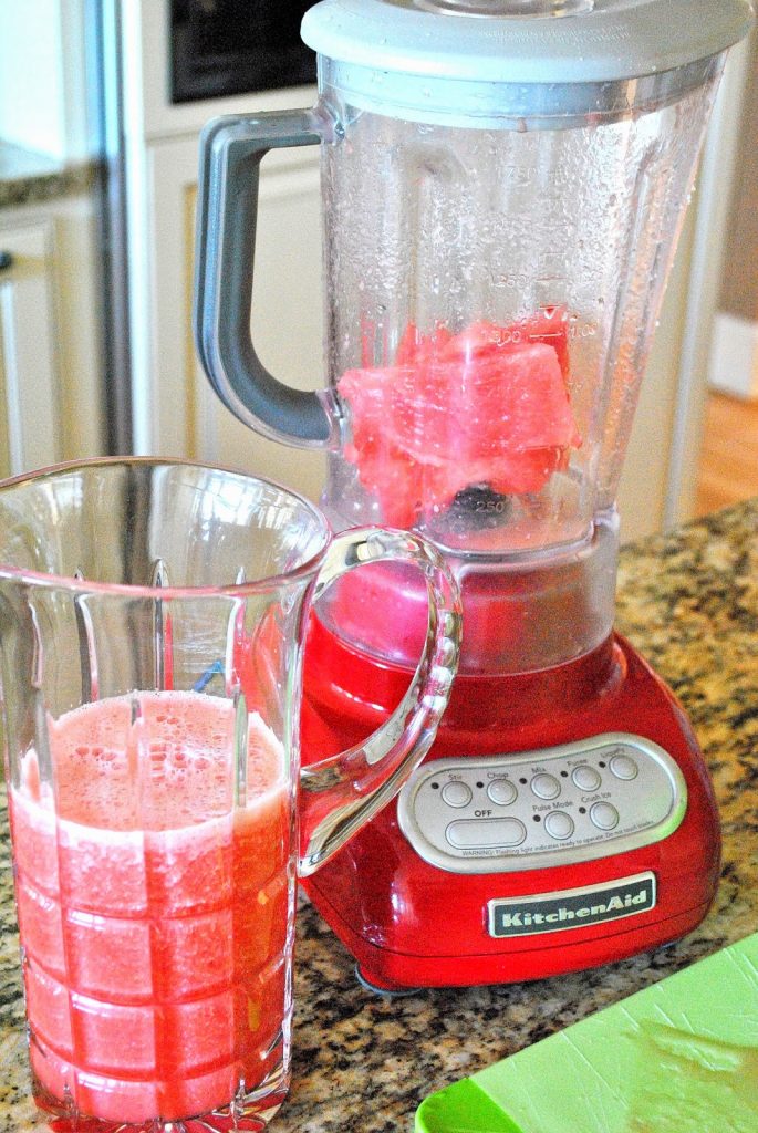 A tall glass pitcher filled with watermelon lemonade and a blender with chunks of watermelon in it. 