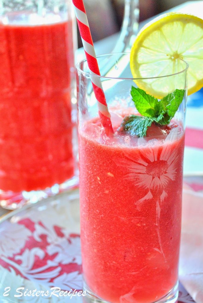 A tall glass filled with watermelon lemonade, a straw, slice of lemon and fresh mint in the glass. by 2sistersrecipes.com