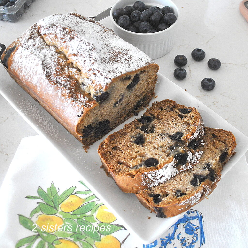 A white platter with a blueberry banana bread sliced with fresh blueberries on the side.