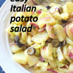 A white round bowl filled with a potato salad, sliced potatoes, olives and celery and capers.