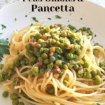 A white dinner plate filled with cooked spaghetti topped with peas and pancetta.