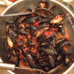 A large silver serving bowl filled with steamed mussels in a tomato based broth and 2 large serving spoons.