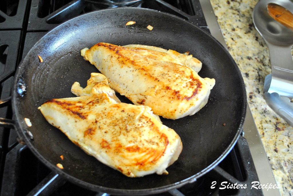 A black skillet on stove top with 2 chicken breasts cooking.