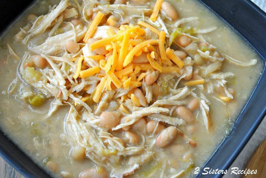 A square bowl filled with white beans, shredded chicken and topped with shredded cheddar cheese.