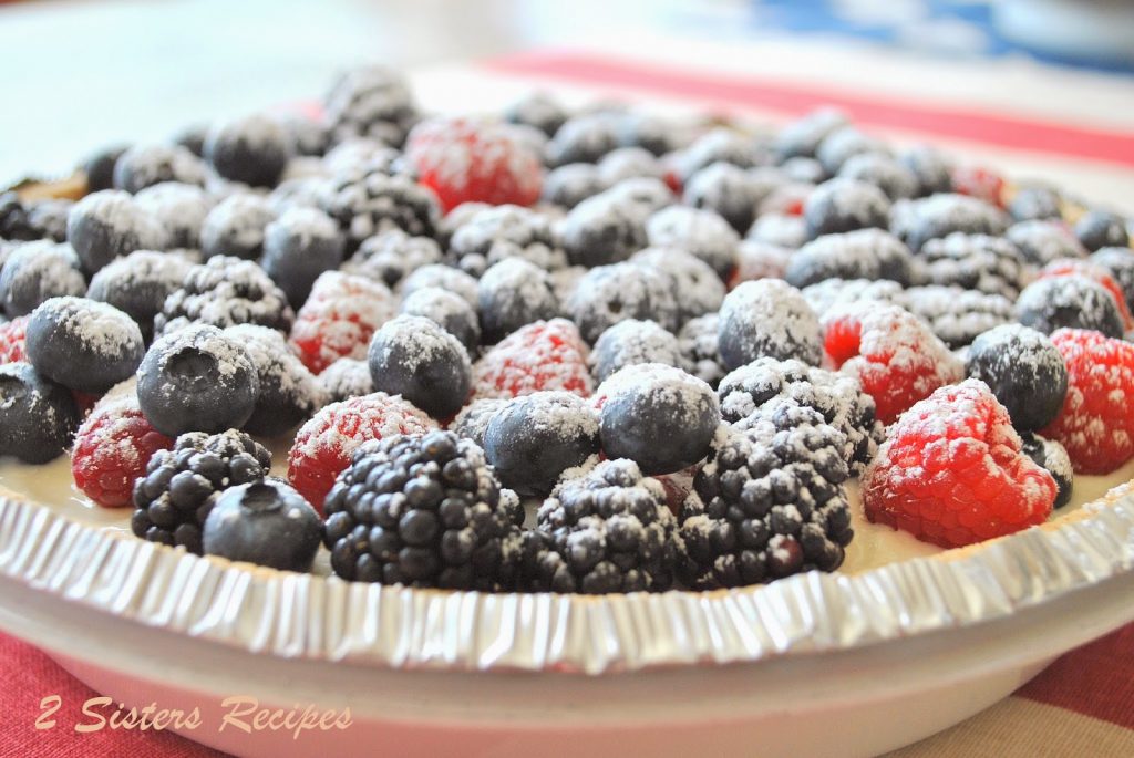 A pie served on the table and topped with mixed fresh berries, and powdered sugar.