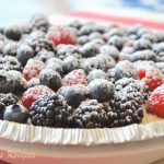 A pie served on the table and topped with mixed fresh berries, and powdered sugar.