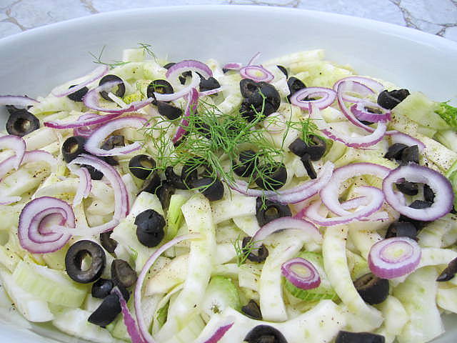 A white oval salad bowl with sliced fennel, red onions, black olives and some fronds tossed on top. 