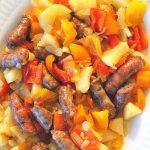 A white platter filled with baked thin sausages, potatoes an colorful peppers.