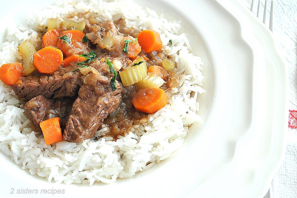 A white plate with cooked rice under a beef stew with carrots and potatoes.