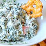 A white bowl filled with a creamy white and green spinach dip with a thin pretzel on the side.
