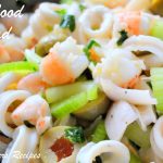 A silver bowl filled with a seafood salad mixed with chopped celery, olives, shrimps, and calamari.