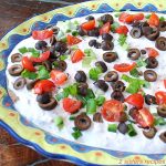 A colorful oval platter filled with layers of creamy topped with chopped green onions, sliced black olives and cherry tomatoes.