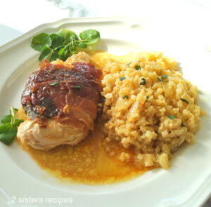 A white dinner plate with a grilled chicken wrapped with crispy prosciutto and a side of cauliflower rice.