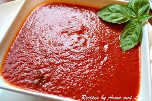 A white square bowl filled with red tomato sauce, and basil leaves on the side.