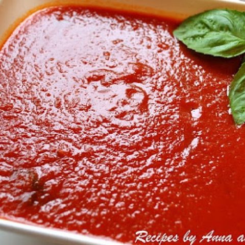A white square bowl filled with red tomato sauce, and basil leaves on the side.