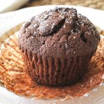 One chocolate muffin with it's paper lining peeled back and served on a white plate.