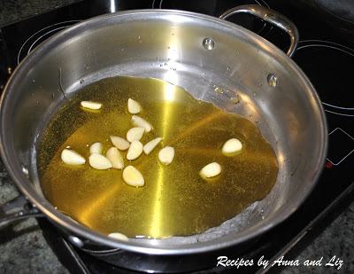A large deep silver skillet on stovetop with olive oil and garlic cloves.