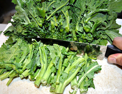 A large cutting knife slicing the ends off of the bunch of broccoli rabe on a cutting board.