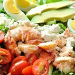 A platter filled with a bed of lettuce, topped with sliced tomatoes, eggs, avocado, and chunks of lobster.