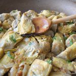 A wooden spoon with an artichoke heart, is scooped up from the skillet.