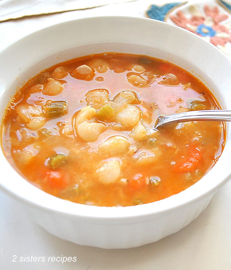 A white bowl filled with beans and vegetables soup with a spoon inside the soup.