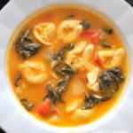 A white soup bowl filled with cheese tortellini, tomato broth and Swiss chard.