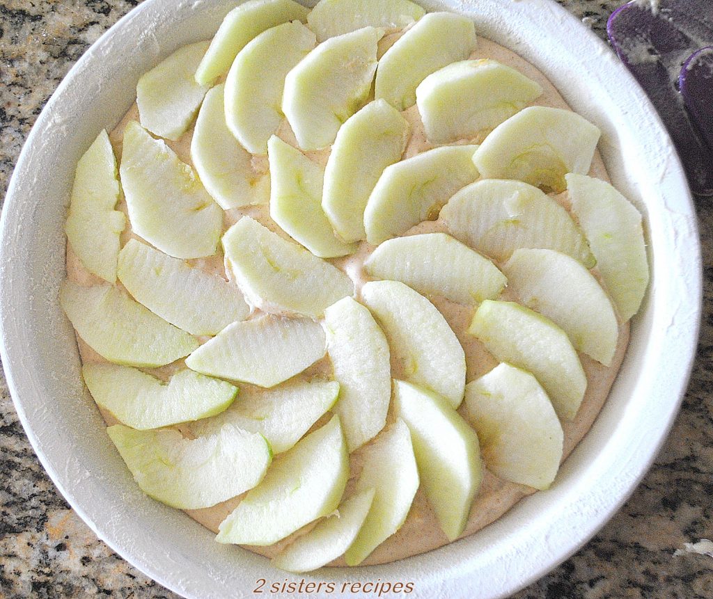  A white pie baking dish with sliced apples covering the batter. by 2sistersrecipes.com