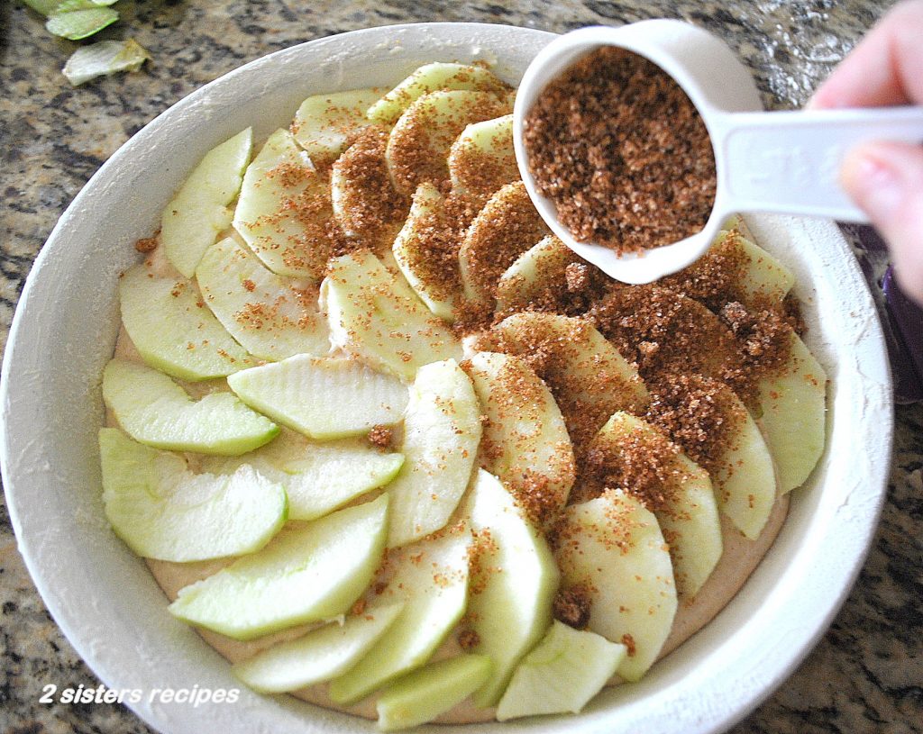 Scattering cinnamon mixture over the sliced apples in before its baked. by 2sistersrecipes.com
