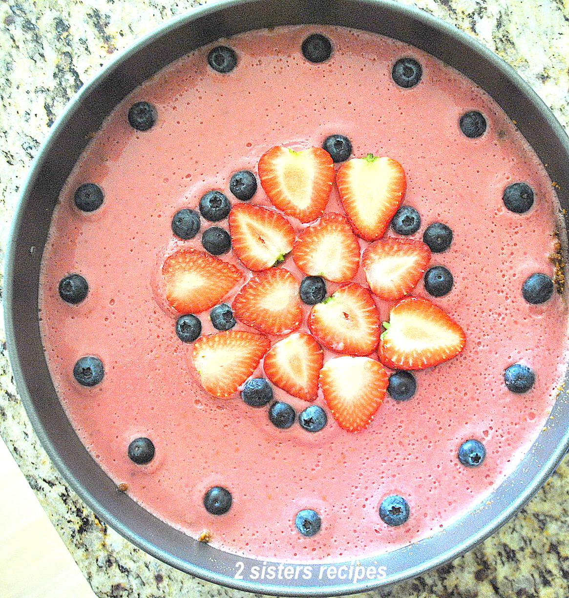 Lemon Raspberry Panna Cotta Cheesecake A springform cake pan with a pink colored top and blueberries with sliced strawberries on top. by 2sisterserecipes.com