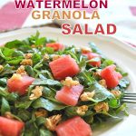 A white plate filled with arugula lettuce, bites of watermelon and granola scattered on top.