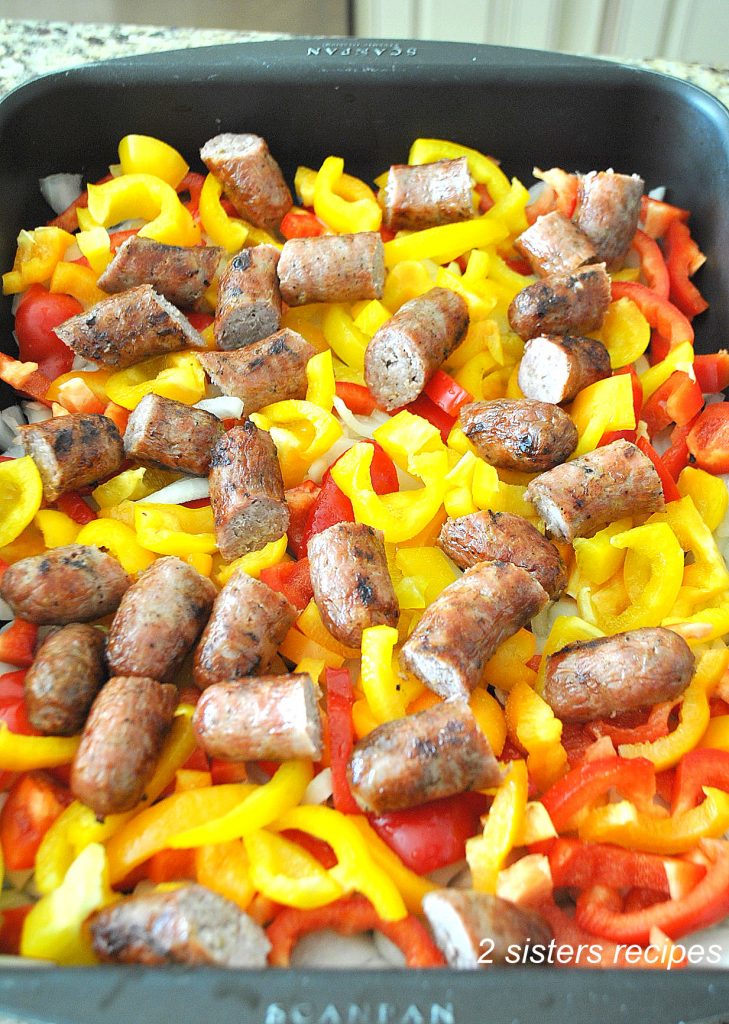 A large baking pan with uncooked peppers and onions, and partially cooked sausages links.