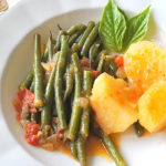 A white plate filled with cooked string beans, and potatoes with fresh basil leaves on the side.