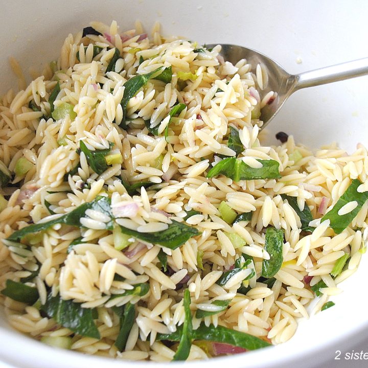 A large white salad bowl with a serving spoon inside, orzo pasta salad with ribbons of spinach and chopped olives.