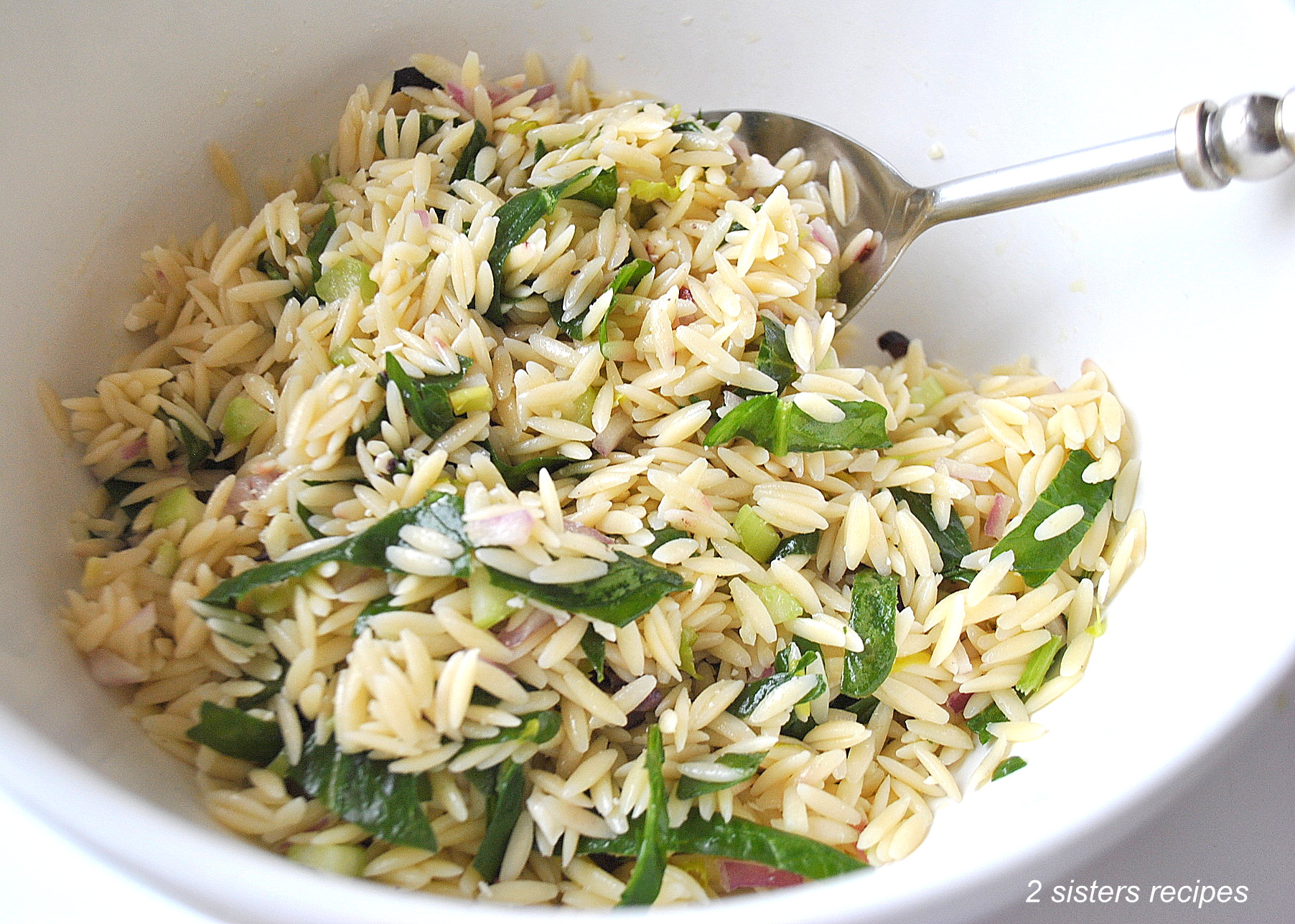 A large white salad bowl with a serving spoon inside, orzo pasta salad with ribbons of spinach and chopped olives.