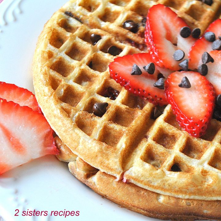 On a white plate, round shape waffles with sliced strawberries and mini chocolate chips on top. by 2sistersrecipes.com