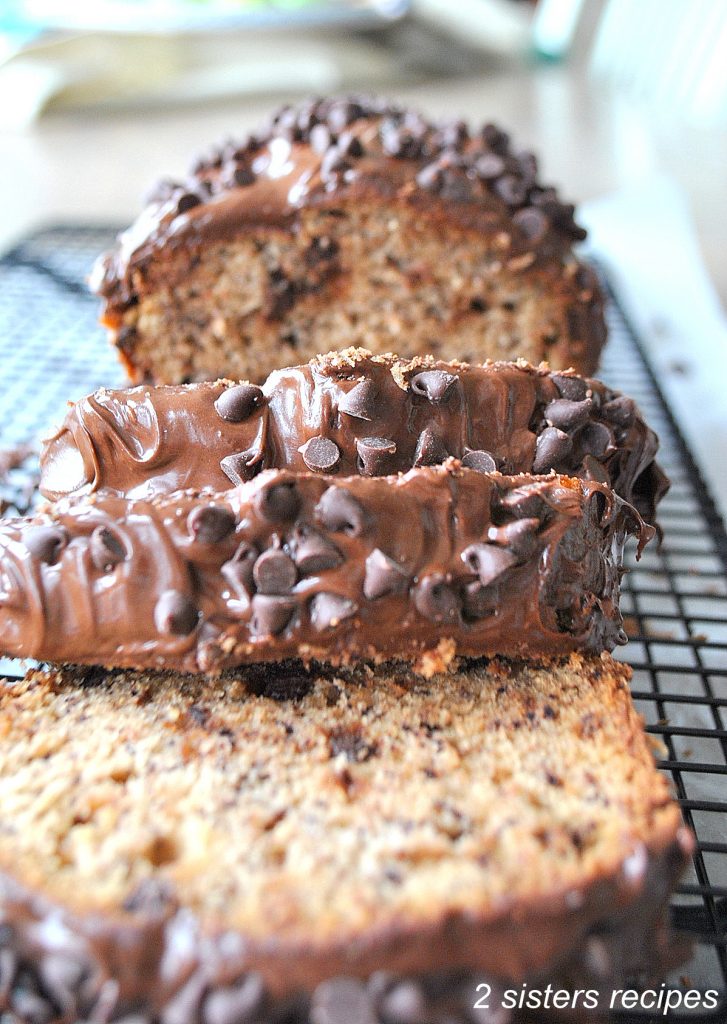 A banana bread with chocolate frosting and chocolate chips sliced on a wire rack.